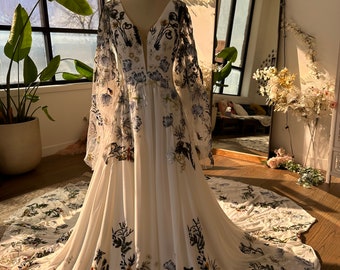 Pode incluir: Um vestido de noiva branco com decote profundo e mangas compridas. O vestido é coberto por um padrão floral com detalhes em preto, azul e verde. O vestido tem uma saia fluida e uma cauda.
