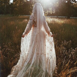 May include: A woman in a white, flowing gown with a veil and star-shaped embroidery walks through a field of tall grass.