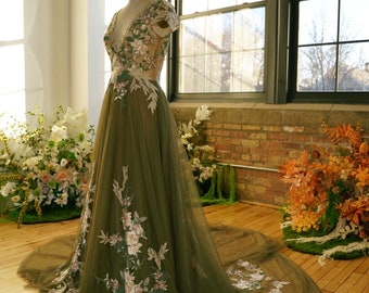 Pode incluir: Um vestido de noiva de tule verde com bordados florais. O vestido tem um decote em V e mangas curtas. O vestido é exibido em um manequim em um estúdio.