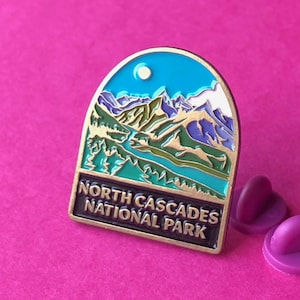 May include: Enamel pin featuring a mountain landscape with a river and the text "North Cascades National Park." The pin has a blue sky, green and purple mountains, and a gold border. Two purple pin backs are included.