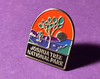 May include: A black and gold enamel pin with a colorful image of a Joshua Tree and the text "Joshua Tree National Park".