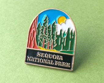 May include: A colorful enamel pin featuring the text "SEQUOIA NATIONAL PARK." The pin depicts a mountain scene with trees, a sun, and a blue sky. The pin has a gold border and is set against a green background.