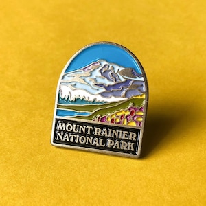 May include: A metal pin with a blue, white, and green enamel design of Mount Rainier National Park. The text "Mount Rainier National Park" is printed in black on the bottom of the pin.