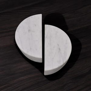 May include: Two white marble half-circle shapes, positioned to form a heart-like design. The marble has subtle gray veining. The objects are set against a dark wood grain background, creating a contrast in textures and tones.