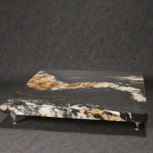 May include: A rectangular black granite serving tray with a natural edge, featuring streaks of gold and white. The tray is supported by four silver-colored metal feet, adding a touch of elegance to the design.