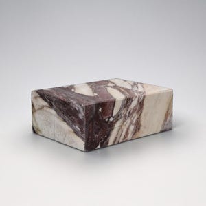 May include: A rectangular block of marble with a striking pattern of burgundy and cream veining. The stone has a polished surface and a smooth, geometric shape. The block is a decorative item.