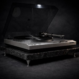 May include: A silver turntable with a black vinyl record playing, set on a black and gray marble stand. The turntable's dust cover is open. The stand has silver cone-shaped feet. The background is dark.