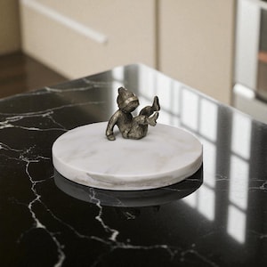May include: A small, bronze-colored figurine of a person in a seated position, wearing a hat, atop a round, white marble tray. The tray is set on a black, veined surface. The figurine is approximately 3 inches tall.