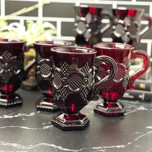 Pode incluir: Conjunto de seis canecas de vidro vermelho com um padrão geométrico em relevo. As canecas têm alças e uma base com pé.