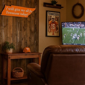 May include: A brown leather recliner facing a television displaying a football game. A wooden console table holds a small potted plant, an orange baseball cap with a white T logo, and a woven basket. A Tennessee sign and poster hang on the wall.
