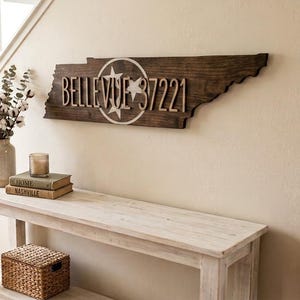 May include: A dark brown wooden wall sign shaped like the state of Tennessee, featuring a white circular emblem with three stars and the text "BELLEVUE 37221" in raised, light-colored lettering mounted on the surface of the wooden state cutout.
