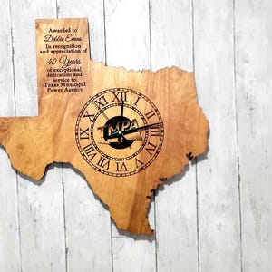 May include: Wooden Texas-shaped clock with Roman numerals and clock hands. The clock face features the letters "MPA" and is engraved with text. The clock is mounted on a light-colored, vertical-planked wall.