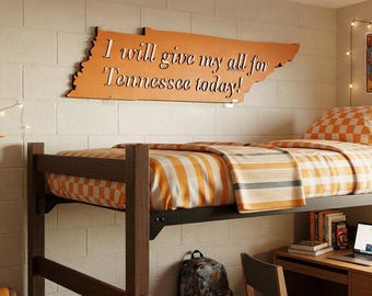May include: A lofted bed with orange and white checkered bedding, featuring a wooden frame. Above the bed, an orange wall sign shaped like Tennessee displays the text I will give my all for Tennessee today! A pennant and posters decorate the wall.