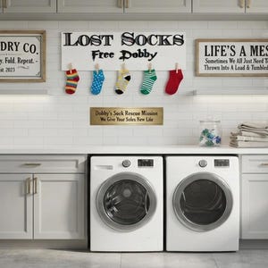 May include: A white wooden sign with the black text LOST SOCKS Free Dobby, featuring five colorful socks clipped to the bottom. Above the white washing machines are two additional framed signs with black text and a small rectangular sign below.