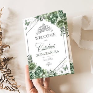 May include: A white card with a silver glitter background and a silver hexagonal frame. The card has a green floral design with a silver crown and the text "Welcome to Catalina's Quinceañera 06.15.20XX".
