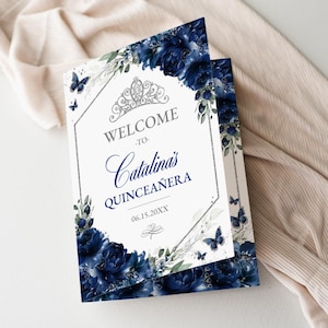 May include: A folded invitation with a white background, navy blue floral accents, and butterflies. The text reads "WELCOME to Catalina's QUINCEAÑERA" with the date 06.15.20XX. A silver tiara and decorative border complete the design.