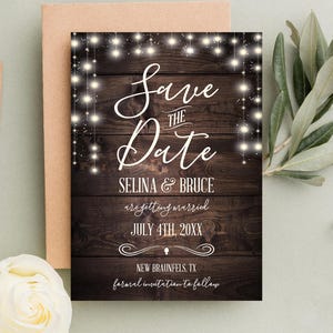 Könnte beinhalten: Rustikale Save-the-Date-Karte mit Holzmaserung und weißen Lichterketten. Der Text lautet "Save the Date" mit den Namen des Paares, dem Hochzeitsdatum und dem Ort.
