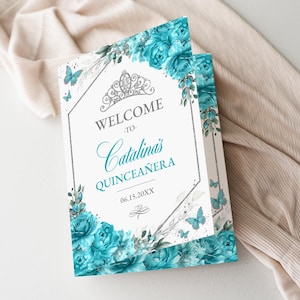 May include: A folded invitation with a floral design. The invitation features the text "WELCOME to Catalina's QUINCEAÑERA" in teal script, with a silver tiara and butterfly accents. Teal flowers and foliage frame the text.