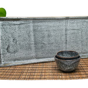 May include: A gray rectangular ceramic serving tray with a speckled glaze. Two gray ceramic bowls are stacked on top of each other on a woven bamboo mat.