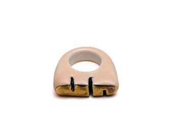 May include: A ceramic ring with a gold and black design. The ring is a light pink color and has a rounded shape.