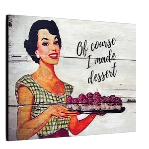 May include: A rectangular wall art piece featuring a retro-style illustration of a person with dark hair wearing a green and white checkered top. The person holds a tray of pink cupcakes. The text reads, Of course I made dessert.