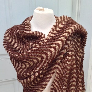 May include: A brown and beige knitted shawl with a chevron pattern. The shawl is draped over a white mannequin, showcasing the intricate design and texture of the handmade garment. The color palette is neutral, making it a versatile accessory.
