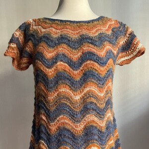 May include: A short-sleeved, hand-knitted top with a chevron pattern in shades of orange, blue, and brown. The top has a scoop neckline and a slightly flared hem. The sleeves are edged with a contrasting white yarn.
