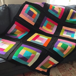 Puede incluir: Una manta tejida que presenta un patr&oacute;n de caba&ntilde;a de troncos compuesto por m&uacute;ltiples bloques cuadrados. Cada bloque contiene tiras rectangulares conc&eacute;ntricas en varios colores brillantes, incluyendo rojo, naranja, amarillo, verde, azul y morado, todos enmarcados por bordes negros.