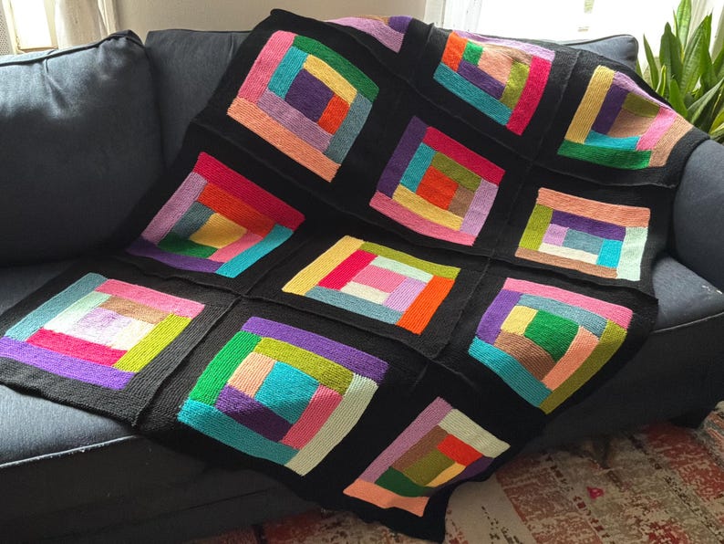 Puede incluir: Una manta tejida que presenta un patr&oacute;n de caba&ntilde;a de troncos compuesto por m&uacute;ltiples bloques cuadrados. Cada bloque contiene tiras rectangulares conc&eacute;ntricas en varios colores brillantes, incluyendo rojo, naranja, amarillo, verde, azul y morado, todos enmarcados por bordes negros.