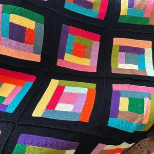 Puede incluir: Una manta tejida que presenta una cuadr&iacute;cula de bloques cuadrados, cada uno compuesto por tiras rectangulares de varios colores dispuestas en un patr&oacute;n de caba&ntilde;a de troncos. Los cuadrados est&aacute;n separados por bordes negros s&oacute;lidos, mostrando una paleta vibrante de hilos rosa, naranja, amarillo, verde, azul y morado.