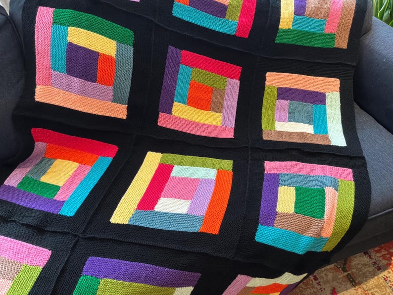 Puede incluir: Una manta tejida que presenta una cuadr&iacute;cula de bloques cuadrados, cada uno compuesto por tiras rectangulares de varios colores dispuestas en un patr&oacute;n de caba&ntilde;a de troncos. Los cuadrados est&aacute;n separados por bordes negros s&oacute;lidos, mostrando una paleta vibrante de hilos rosa, naranja, amarillo, verde, azul y morado.