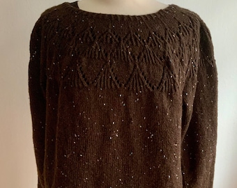 May include: A brown knit sweater with a round neckline and long sleeves. The sweater features a detailed openwork pattern across the upper chest and small white flecks throughout the fabric. The sweater is displayed on a mannequin.