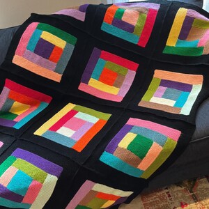 Puede incluir: Una manta tejida que presenta una cuadr&iacute;cula de bloques cuadrados, cada uno compuesto por tiras de lana multicolor dispuestas en un patr&oacute;n de caba&ntilde;a de troncos. Los cuadrados est&aacute;n separados por gruesos bordes negros, mostrando una paleta vibrante de tonos rojos, naranjas, amarillos, verdes, azules, morados y rosas.