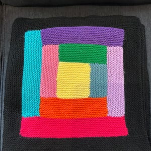 Puede incluir: Una funda de coj&iacute;n tejida cuadrada con un patr&oacute;n de caba&ntilde;a de troncos con un rect&aacute;ngulo central amarillo rodeado por bloques de color verde, azul, naranja, rosa, morado y morado claro, enmarcado por un borde tejido negro s&oacute;lido.