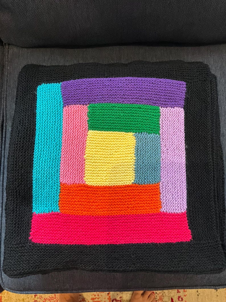 Puede incluir: Una funda de coj&iacute;n tejida cuadrada con un patr&oacute;n de caba&ntilde;a de troncos con un rect&aacute;ngulo central amarillo rodeado por bloques de color verde, azul, naranja, rosa, morado y morado claro, enmarcado por un borde tejido negro s&oacute;lido.