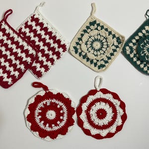 May include: A collection of six handmade crochet pot holders in various designs. Two are red and white zigzag patterns, two are cream and green floral patterns, and two are red and white circular patterns. Each has a loop for hanging.