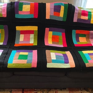 Puede incluir: Una manta tejida a mano que presenta una cuadr&iacute;cula de doce bloques cuadrados. Cada bloque est&aacute; compuesto por tiras de hilo multicolor en tonos rosa, amarillo, naranja, verde, azul y morado, todo enmarcado por un borde negro s&oacute;lido.