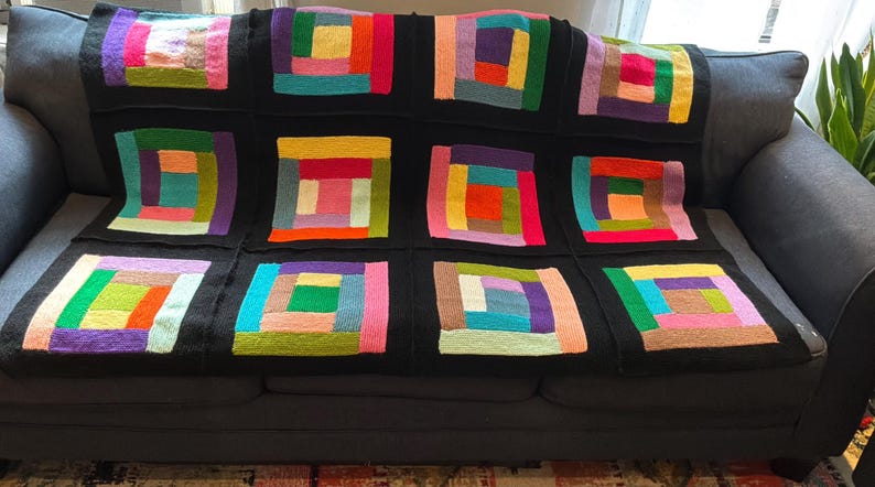 Puede incluir: Una manta tejida a mano que presenta una cuadr&iacute;cula de doce bloques cuadrados. Cada bloque est&aacute; compuesto por tiras de hilo multicolor en tonos rosa, amarillo, naranja, verde, azul y morado, todo enmarcado por un borde negro s&oacute;lido.