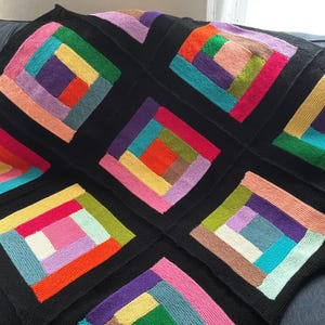 Puede incluir: Una manta tejida que presenta un patr&oacute;n repetitivo de caba&ntilde;a de troncos compuesto por varios bloques cuadrados. Cada bloque consta de tiras rectangulares coloridas en tonos rojo, naranja, amarillo, verde, azul, morado y rosa, enmarcadas por bordes negros.