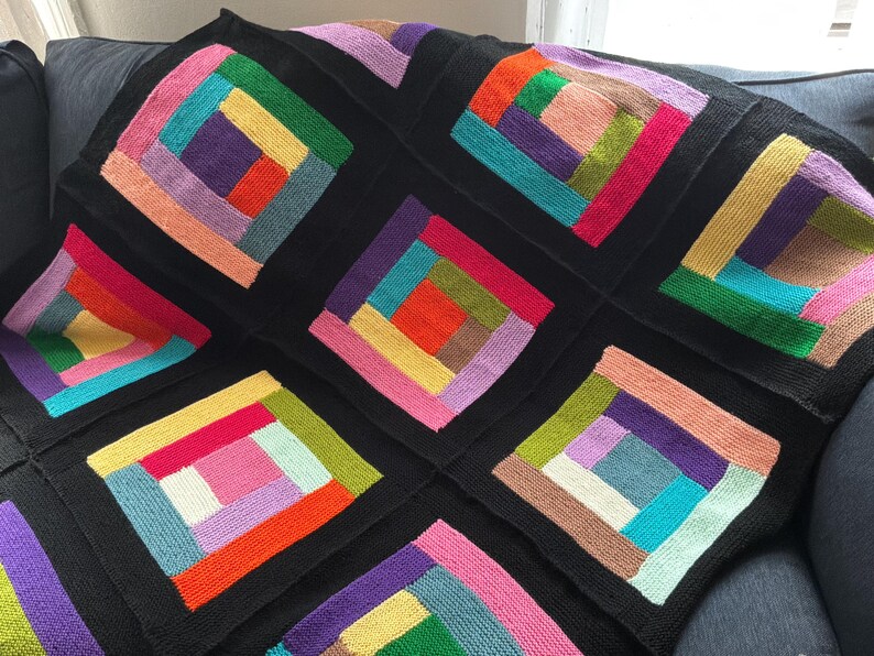 Puede incluir: Una manta tejida que presenta un patr&oacute;n repetitivo de caba&ntilde;a de troncos compuesto por varios bloques cuadrados. Cada bloque consta de tiras rectangulares coloridas en tonos rojo, naranja, amarillo, verde, azul, morado y rosa, enmarcadas por bordes negros.