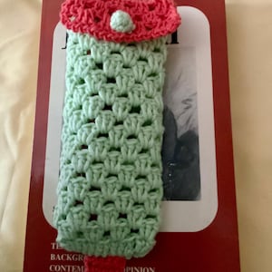 Könnte beinhalten: Handgefertigter Häkel-Lesezeichen in Form eines Eis am Stiel. Das Lesezeichen hat einen hellgrünen Körper, einen roten Stiel und einen roten Deckel mit einem kleinen grünen Knopf. Es liegt auf einem Buch mit rotem Einband.