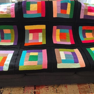 Puede incluir: Una manta tejida que presenta una cuadr&iacute;cula de doce bloques cuadrados, cada uno compuesto por tiras rectangulares de varios colores dispuestas en un patr&oacute;n conc&eacute;ntrico. Los bloques coloridos est&aacute;n separados por secciones negras.