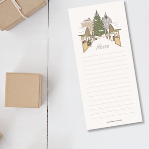 May include: A cream-colored notepad with a winter village illustration and the word "Notes" in script. The notepad has lined pages and a website address at the bottom. Two small, brown gift boxes are in the background.