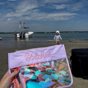 May include: A clear, iridescent pouch with a lavender top and the embroidered word "Stitches" in pink. The pouch is filled with colorful embroidery floss. The background shows a beach scene with a boat and a child.