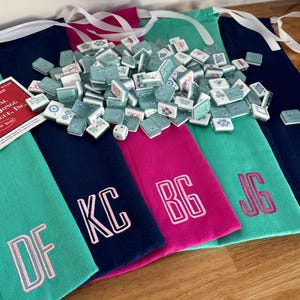 May include: A collection of Mahjong tiles and dice are displayed on top of several colorful fabric pouches. The pouches are teal, navy, and pink, each embroidered with initials. A card with the text "NATIONAL MAH JONGG LEAGUE, INC." is also visible.