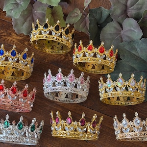 May include: An assortment of ornate crowns in gold, silver, and rose gold tones, adorned with colorful gemstones. The crowns feature various jewel colors, including blue, red, pink, green, and clear, set against a detailed metalwork design.