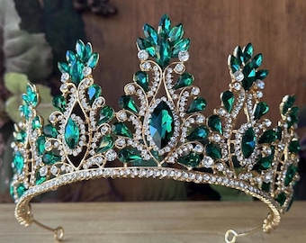 May include: A gold-toned metal crown featuring a series of pointed arches adorned with large, marquise-cut emerald green crystals and smaller, clear round rhinestones. The intricate filigree metalwork is encrusted with clear stones throughout.