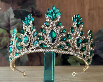 May include: A gold crown with emerald green gemstones and clear rhinestones. The crown is on a stand.