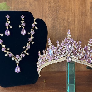 May include: A regal jewelry set and crown. The set includes a necklace and earrings with purple teardrop-shaped gems and floral accents. The gold-toned crown features a similar design with purple and clear crystals. The jewelry is displayed on a black velvet stand.