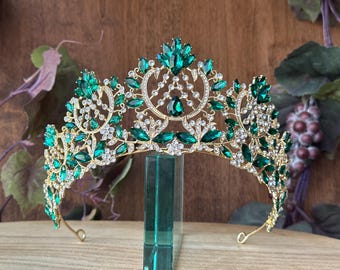 May include: A gold crown with emerald green gemstones and clear rhinestones. The crown is designed with a floral pattern and has a pointed top.